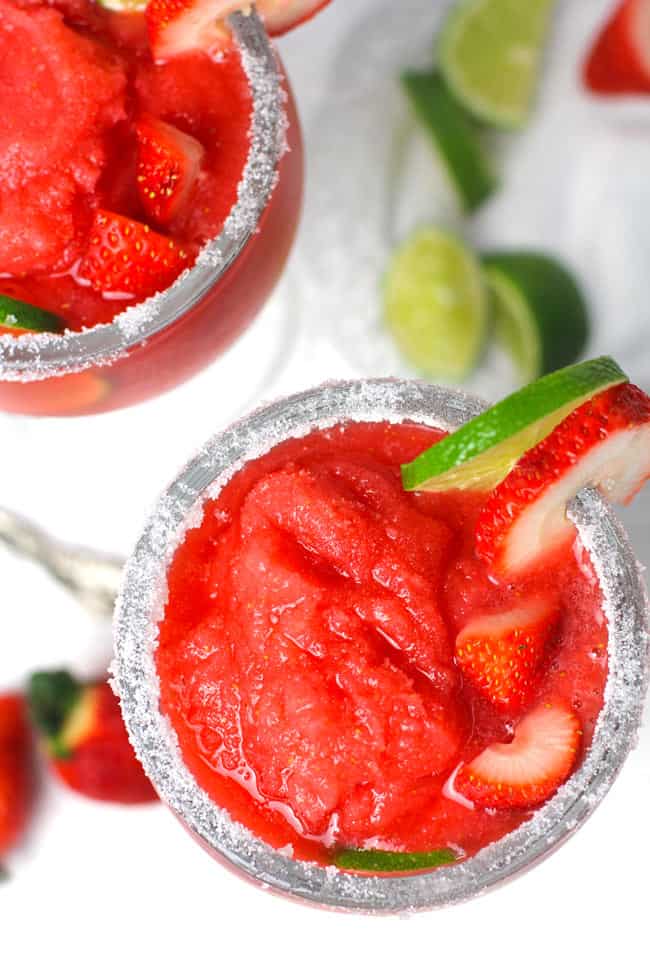 Overhead shot of two frozen strawberry lemonade margaritas, with a salt rim, and lime and strawberry wedges on the glass.