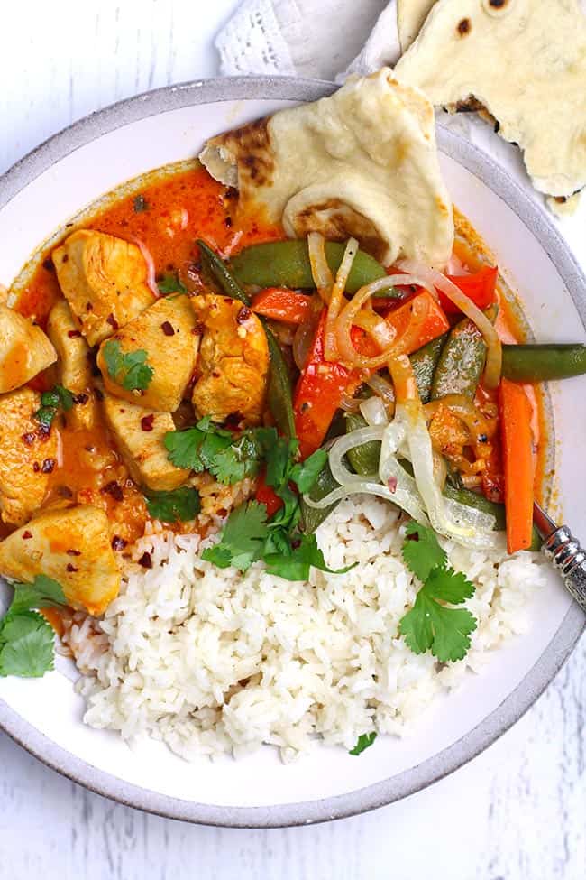 Overhead shot of a white and gray bowl with chicken curry and vegetables, rice, and naan bread.