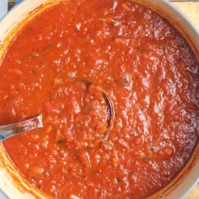Overhead shot of a stock pot of homemade marinara sauce with a ladle inside it, on a wooden board.