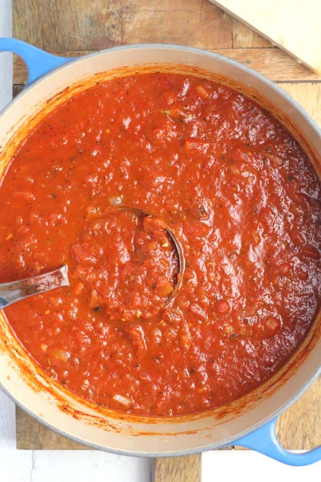 Overhead shot of a stock pot of homemade marinara sauce with a ladle inside it, on a wooden board.