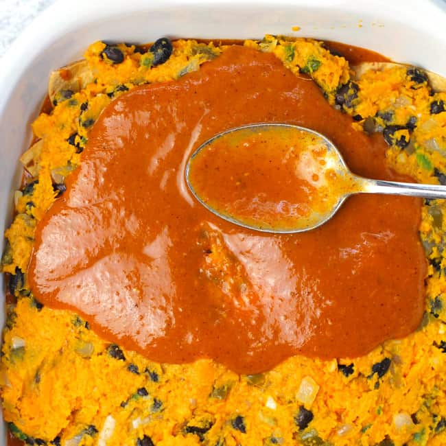Overhead shot of a spoon spreading enchilada sauce over the sweet potato layer.