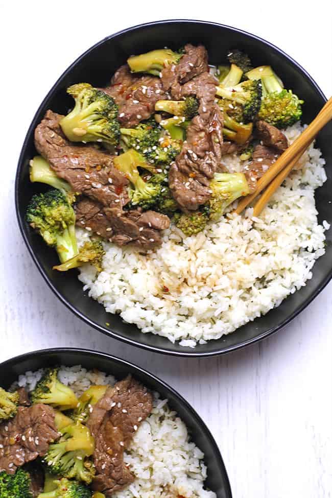 Overhead shot of two black bowls of beef and broccoli stir fry, over rice with chop sticks.