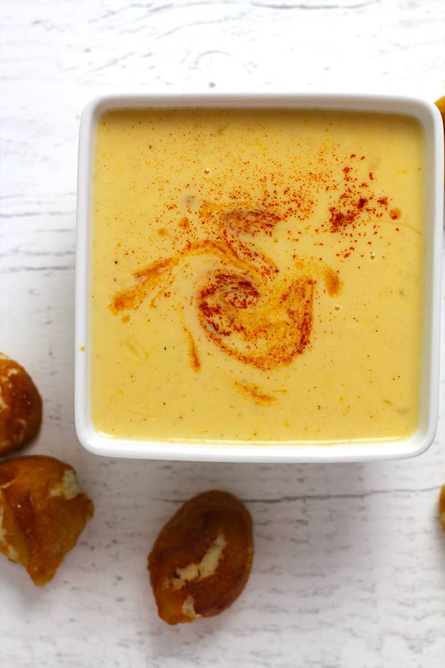 Overhead shot of a square white bowl of spicy beer cheese dip, with pretzel bites next to it.