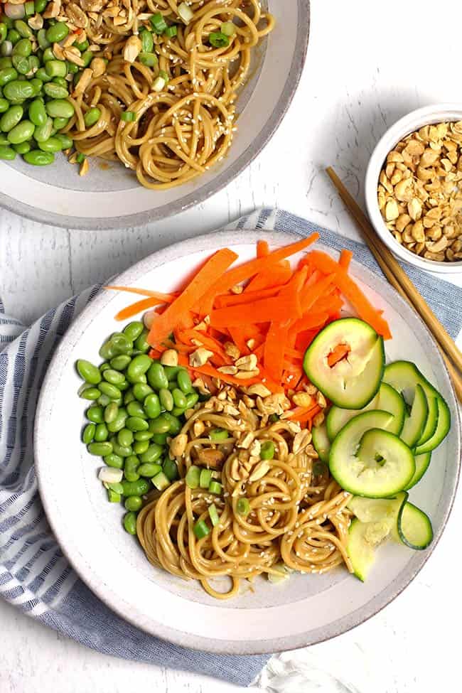 Overhead shot of two bowls of peanut sesame noodles with vegetables, on a white background.