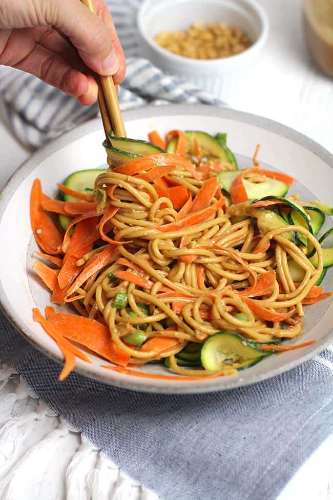Side shot of a white bowl of peanut sesame noodles with vegetables, being twirled with some chopsticks.