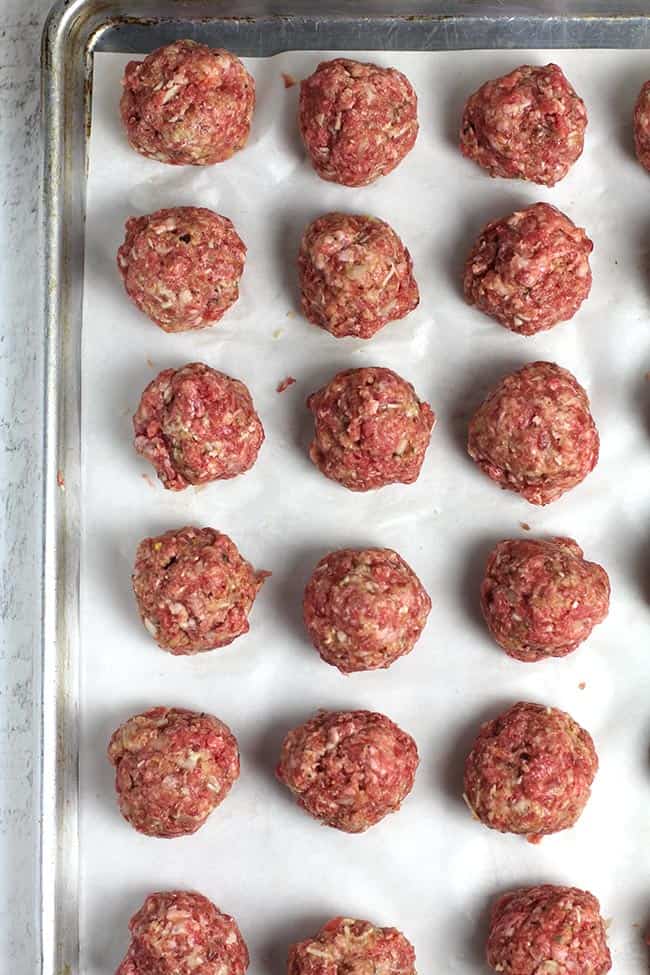 Overhead shot of a baking sheet lined with white parchment paper, with 24 raw meatballs.