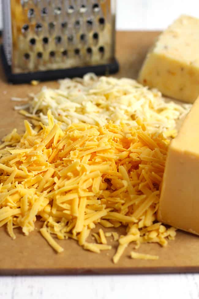 Side shot of a tan board of freshly shredded cheese, next to blocks of cheese.