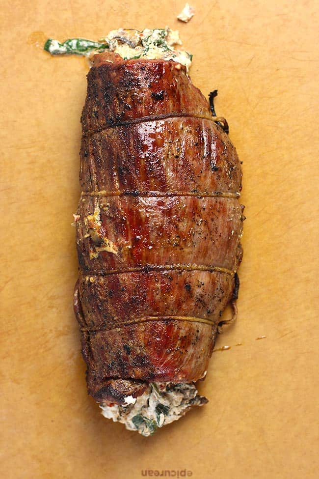 Overhead view of a grilled and stuffed flank steak, tied in a roll with some stuffing falling out.