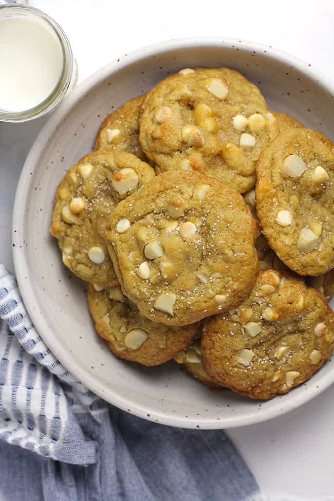 A bowl of white chocolate macadamia nut cookies stacked up, with a jar of milk.