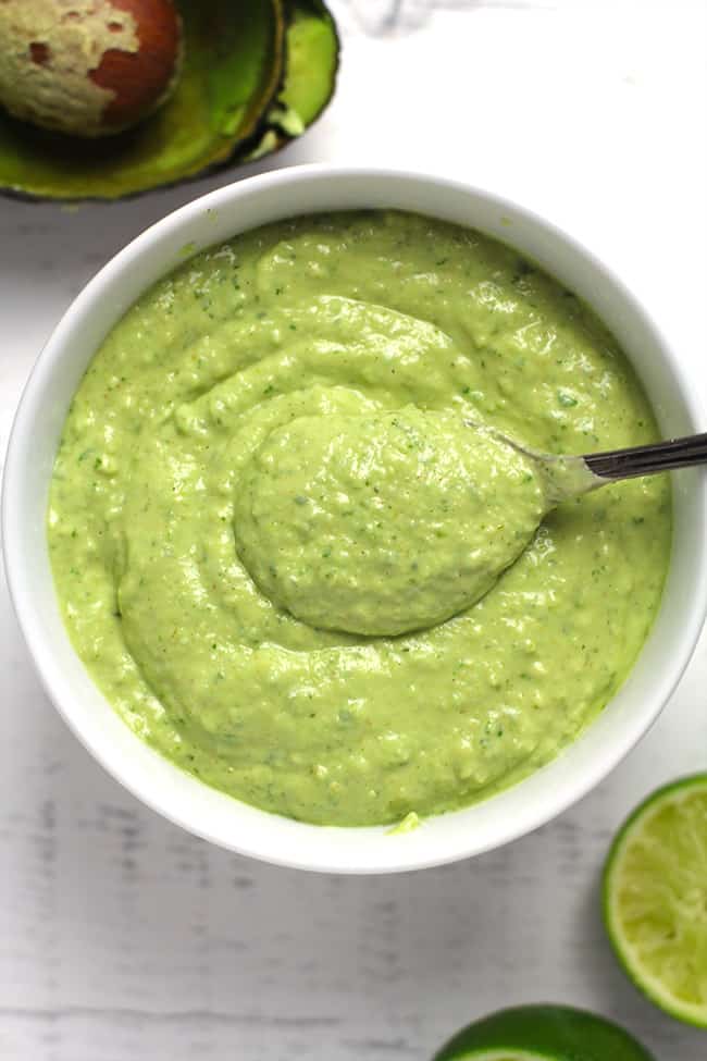 Overhead shot of a white bowl of creamy avocado dressing with a spoon inside, on a white background.