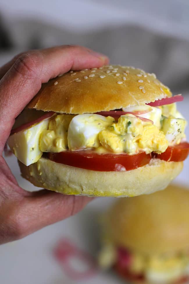 A hand holding a classic egg salad sandwich, showing the creamy insides and tomato slices.
