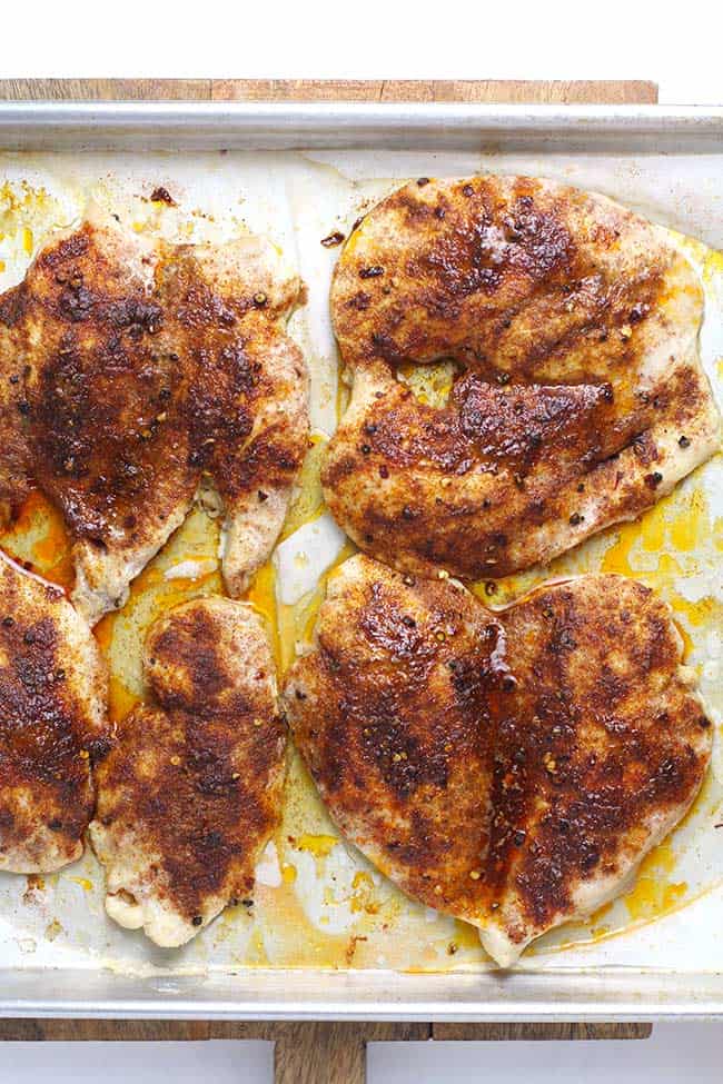 Four butterflied chicken breasts with seasonings and olive oil, on a baking sheet.