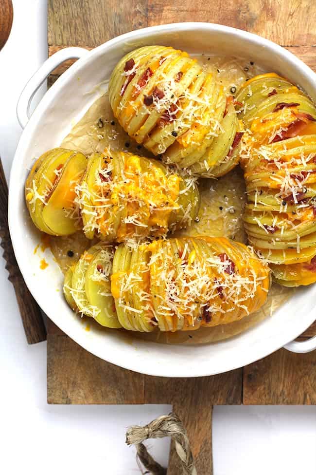 A white dish of four cheesy hasselback potatoes, in a wood board.