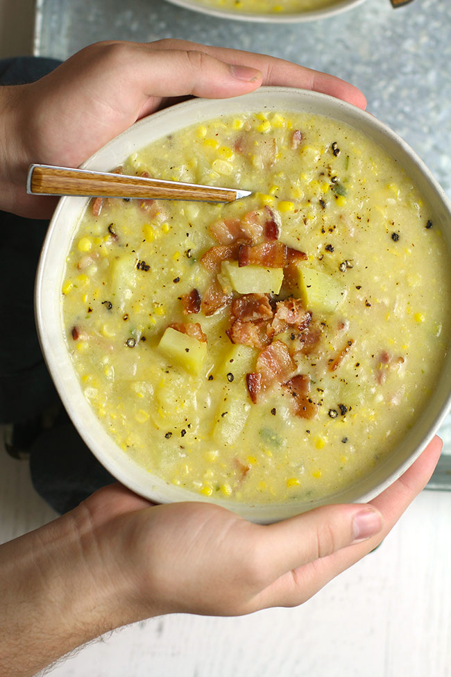 Two hands holding a bowl of corn chowder with bacon.