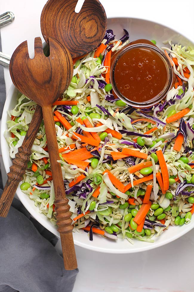 Overhead view of a large white bowl of crunchy asian cabbage salad, with a jar of dressing and wooden spoons.