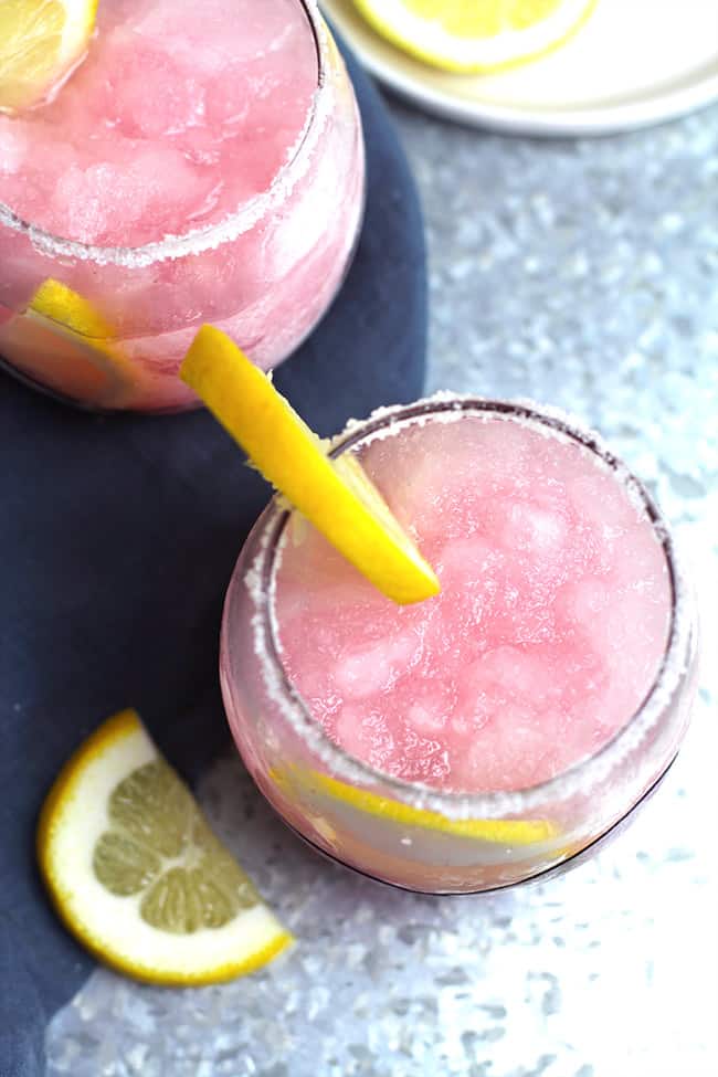 Overhead view of two glasses of pink lemonade vodka slush, with lemon slices, on a gray tray.