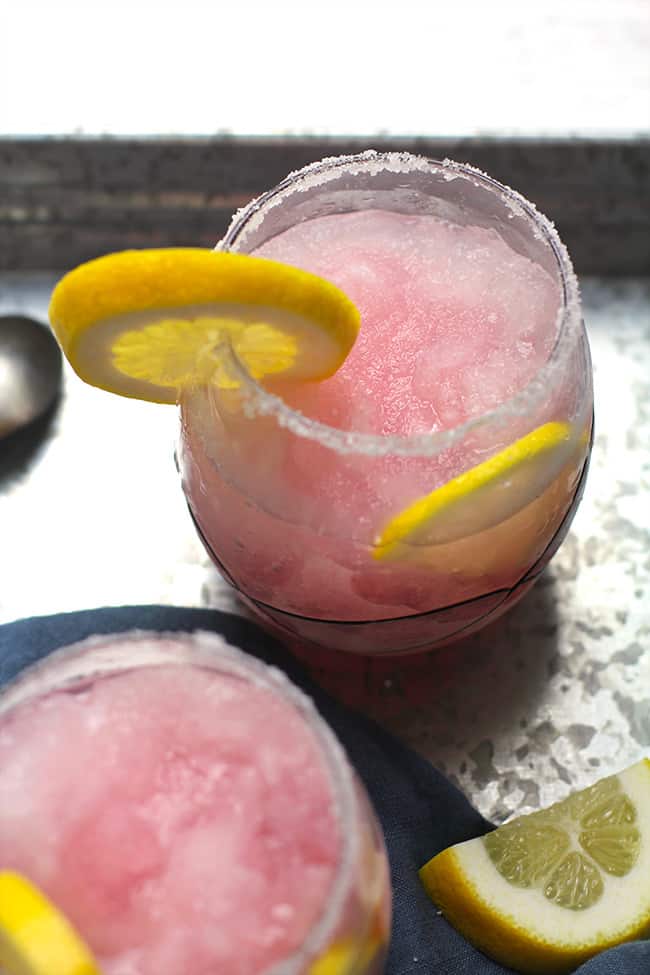 Overhead shot of two glasses of pink lemonade vodka slushes, with lemon wedges.