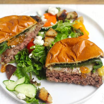 Side shot of a grilled poblano burger with apricot preserves, sliced in half on a white plate with salad.