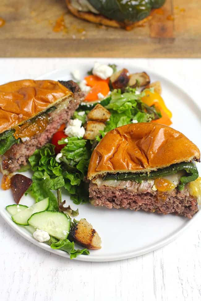 Side shot of a grilled poblano burger with apricot preserves, sliced in half on a white plate with salad.