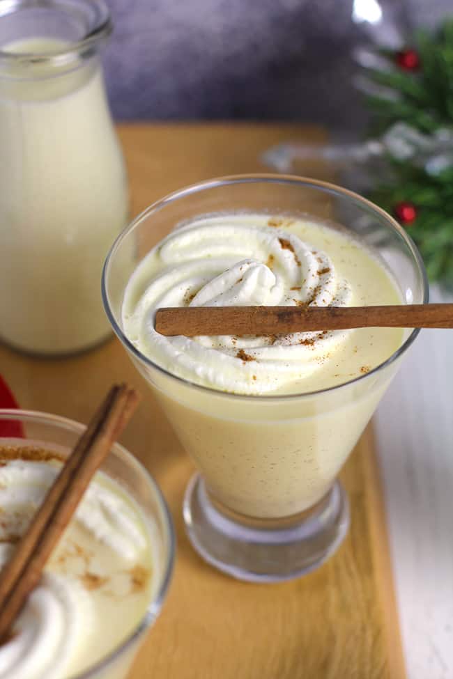 Overhead shot of several glasses of homemade eggnog, with cream and cinnamon sticks on top.