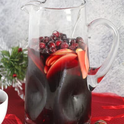 A large pitcher of cranberry sangria recipe, on a red placemat with ornaments around it.
