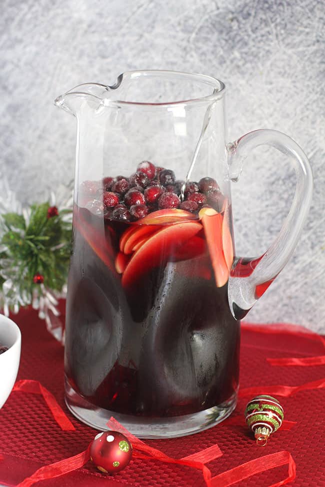 A large pitcher of cranberry sangria recipe, on a red placemat with ornaments around it.