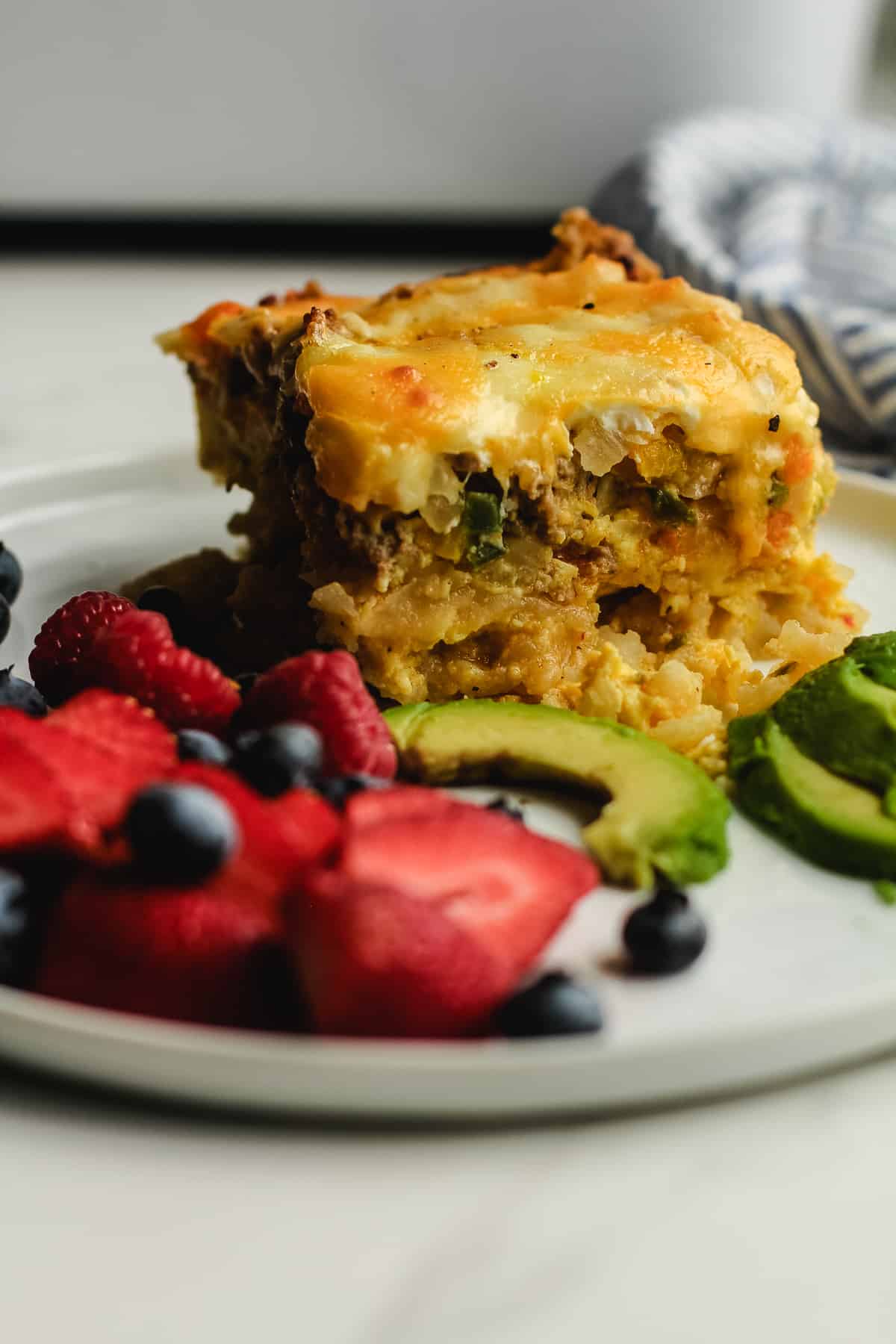 Side shot of a square of breakfast casserole on a plate with avocado and fruit.