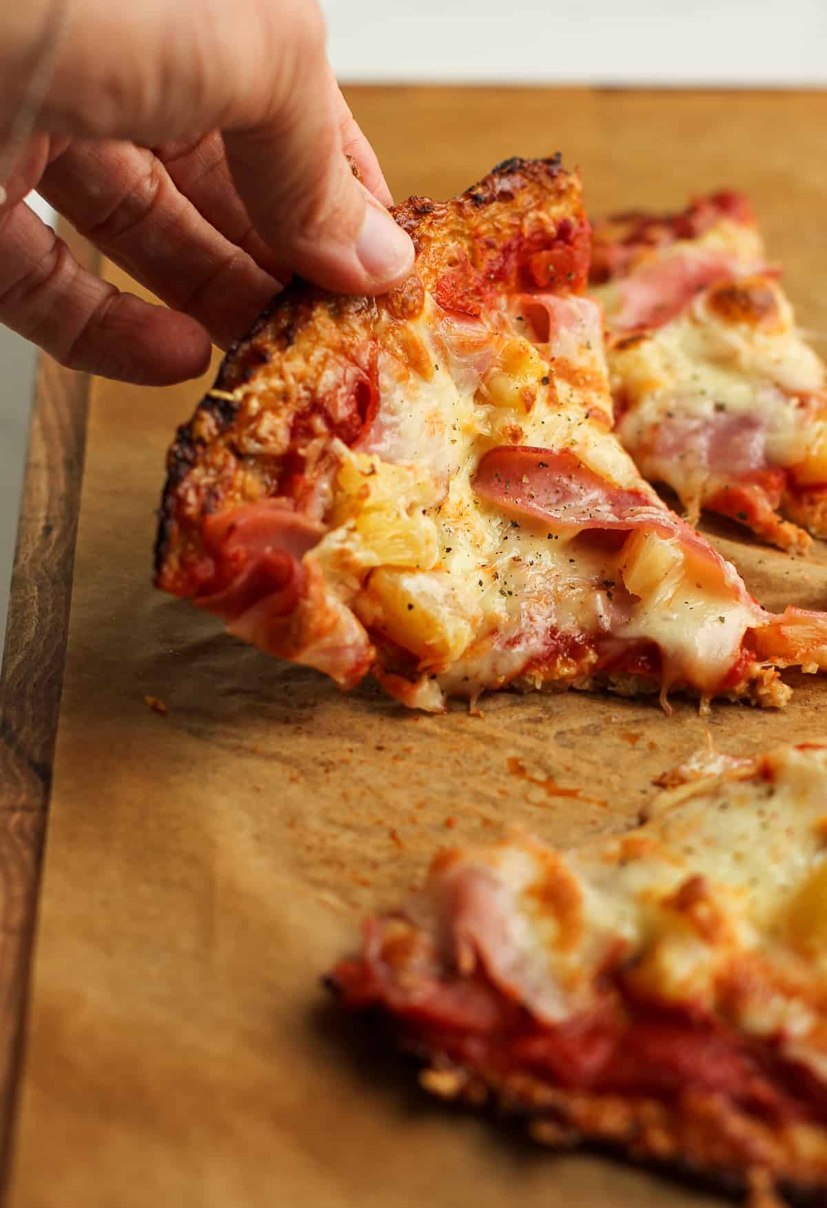 A hand lifting up a piece of Hawaiian pizza on cauliflower crust.