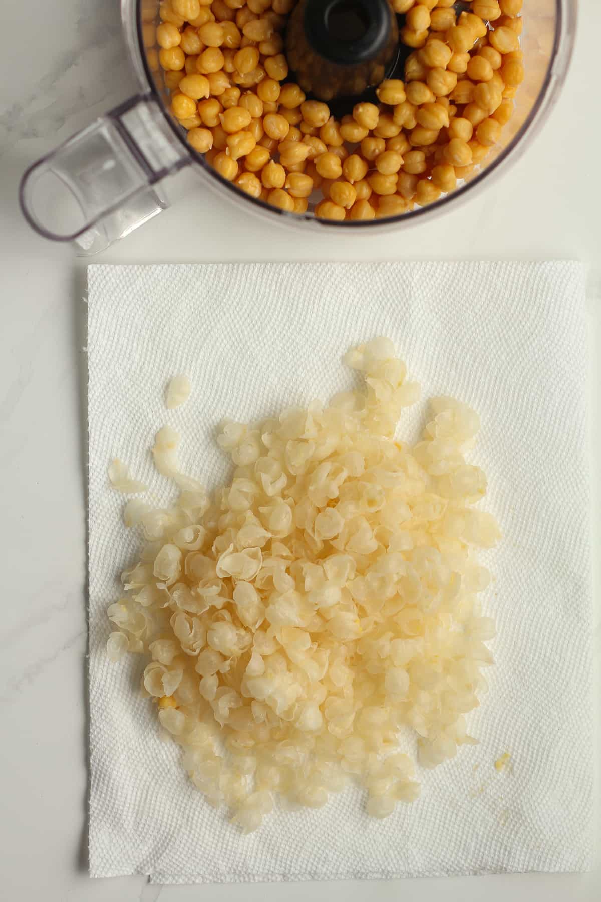 Overhead shot of some chickpea skins next to a food processor of chickpeas.