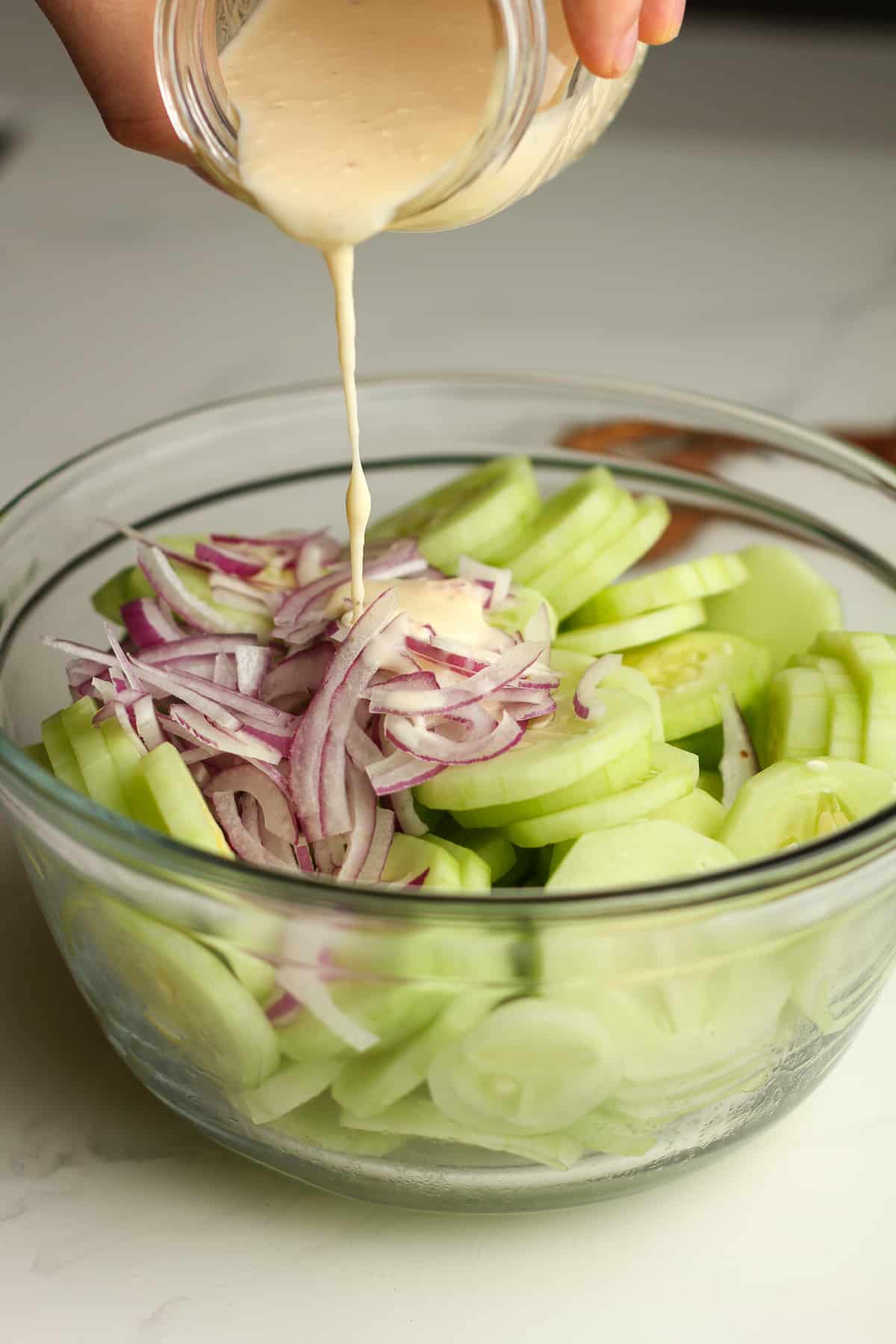 A jar of dressing being poured on a bowl of sliced cucumbers and onions.