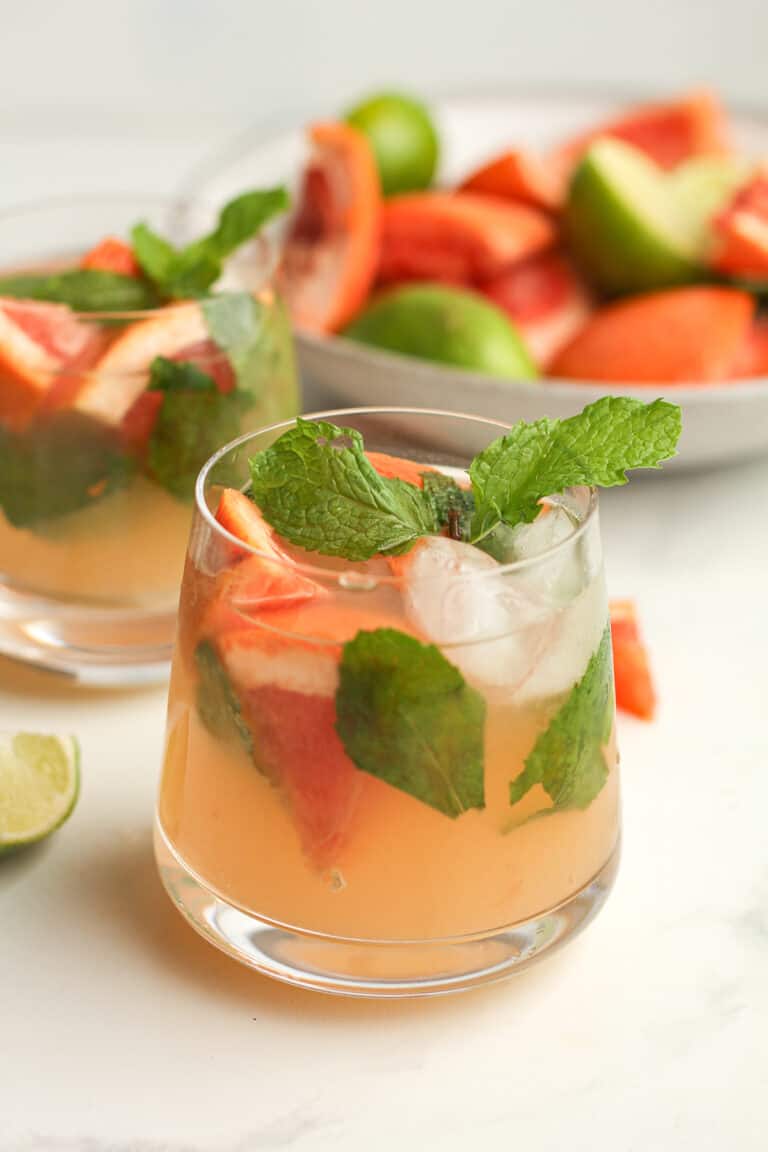 Two glasses of mojitos, with fruit in the background.