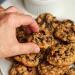 A hand reaching for a mini cranberry oatmeal cookie on a plate of cookies.