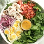 The bowl of spinach salad by ingredient with the jar of dressing.
