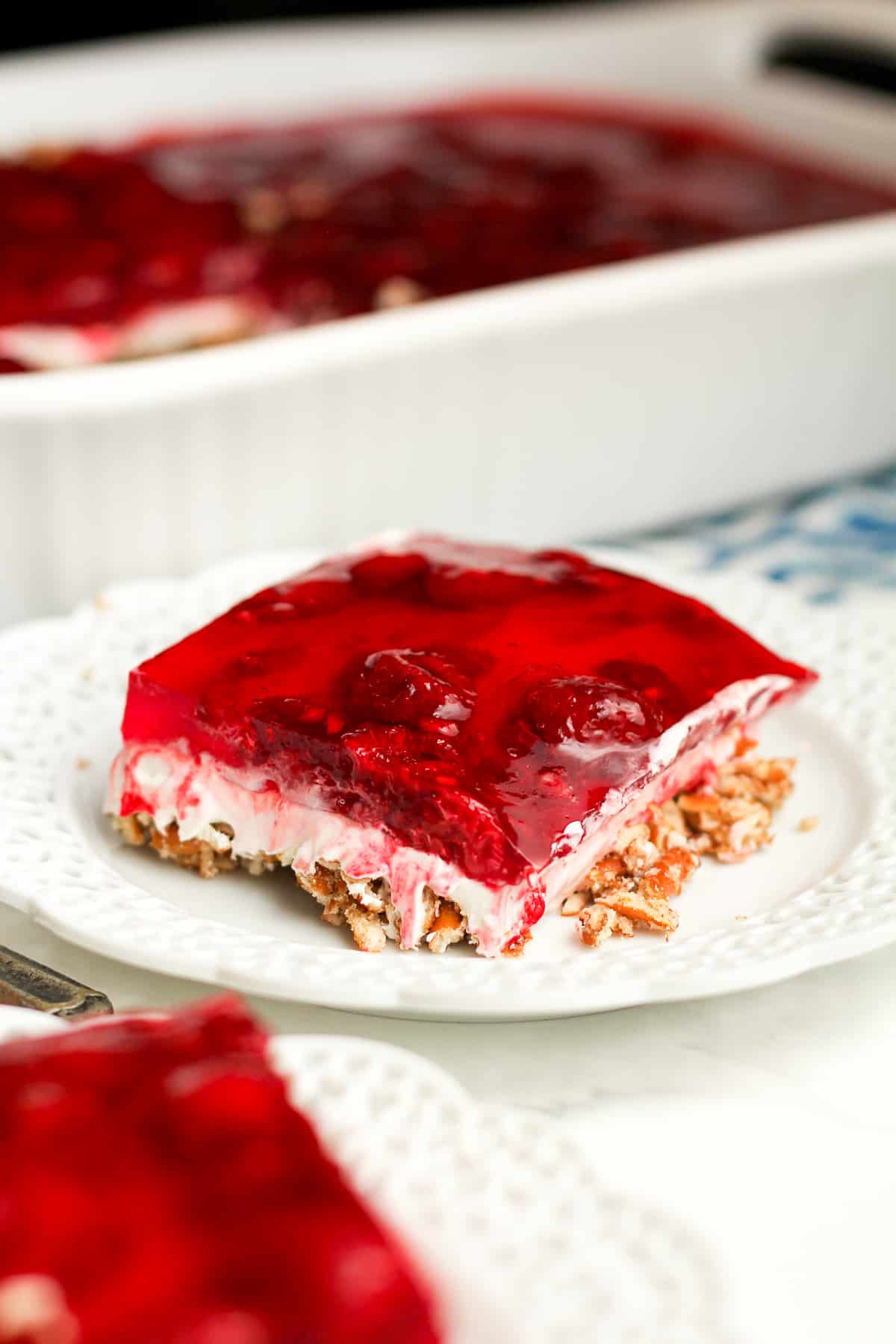 Side shot of a serving of raspberry pretzel salad with the large dish in the back.