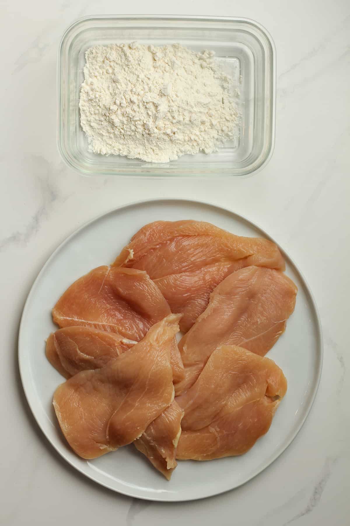 A plate of chicken breasts with a bowl of flour.