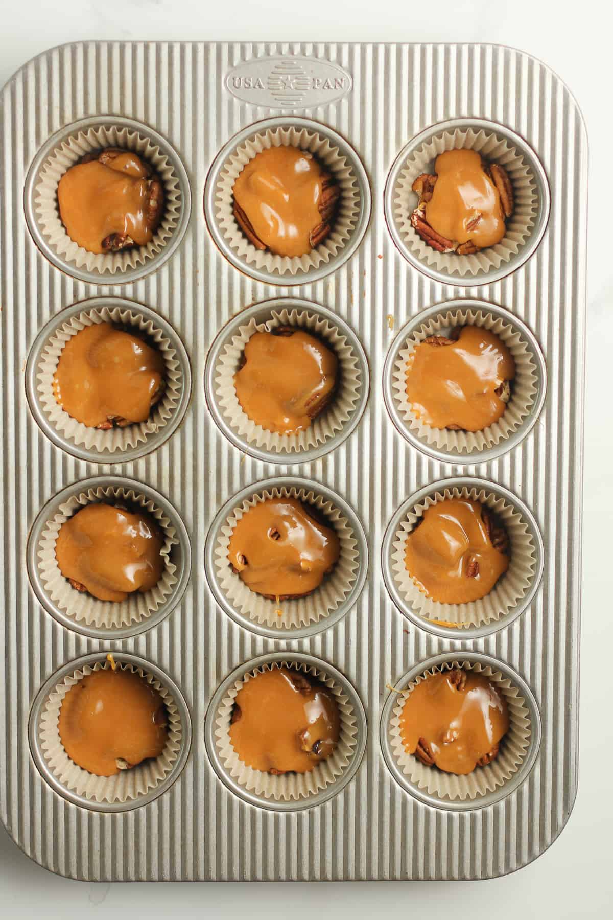A 12-cup muffin tin with caramel on top of pecans in parchment cups.