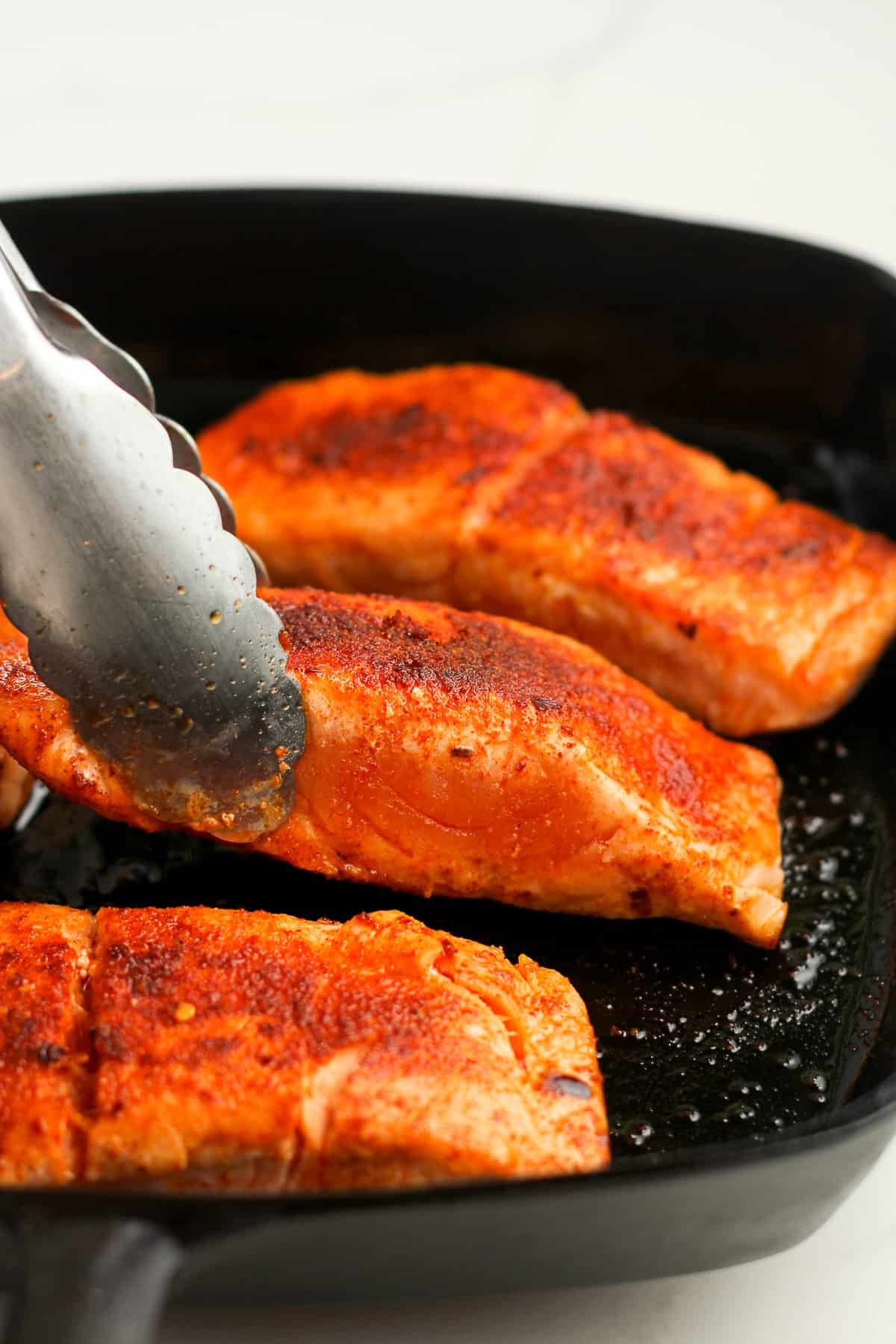 A tongs lifting up a piece of partially cooked blackened salmon.