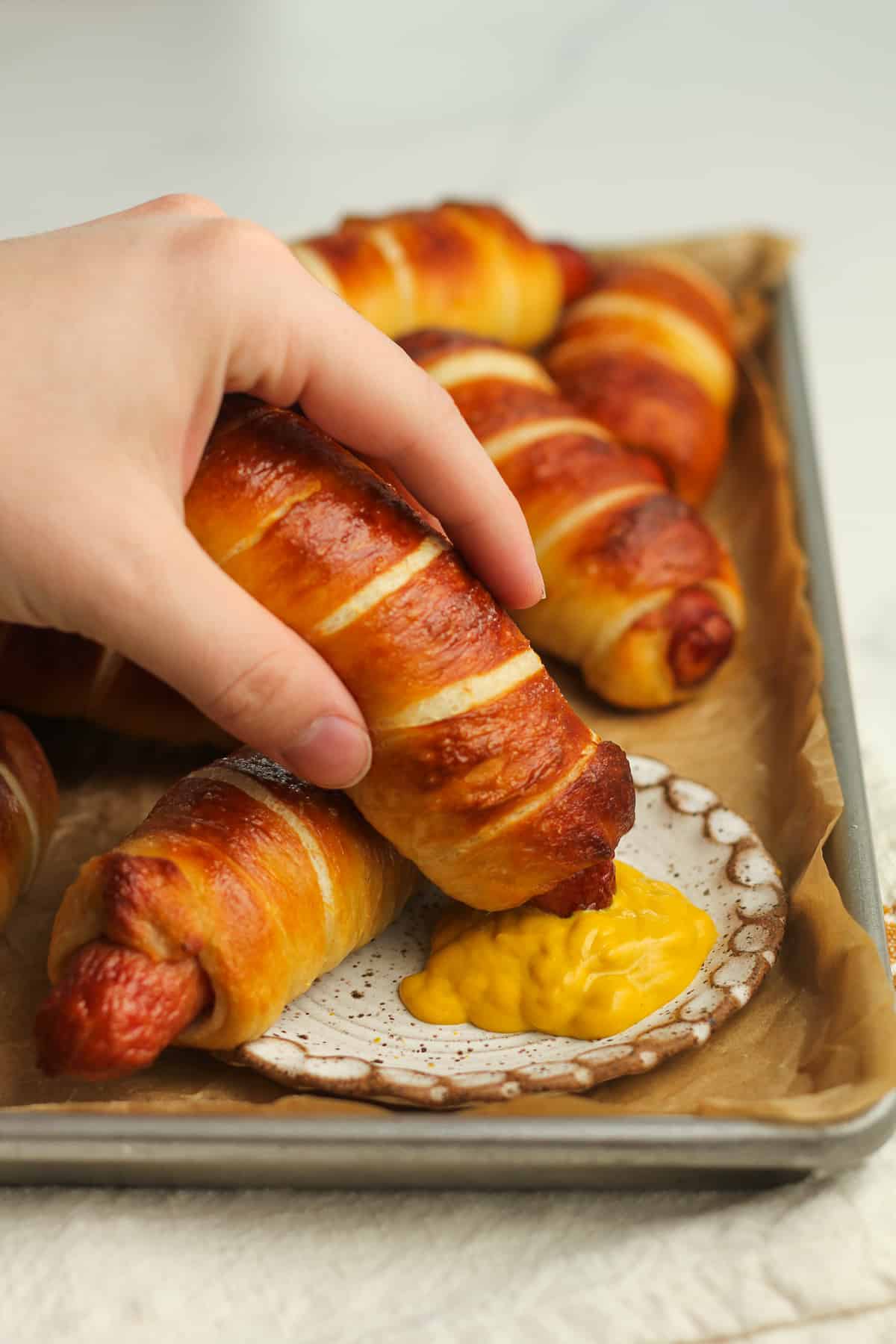 A hand dipping a pretzel dog in some mustard.