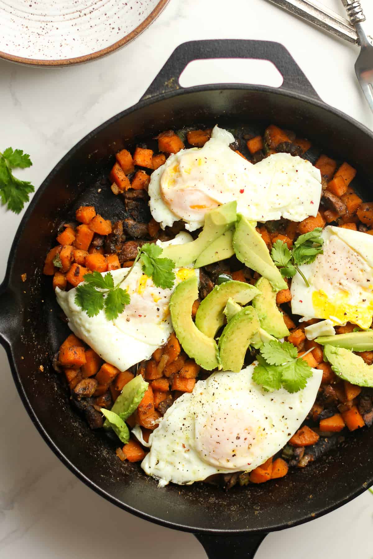 A skillet of the sweet potato hash with the cooked eggs on top with avocado slices.