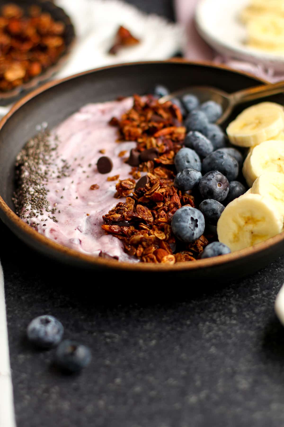 Side shot of a bowl of yogurt, chocolate granola, blueberries, and bananas with chia seeds.