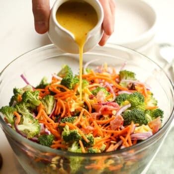 A hand pouring some honey dijon dressing over a broccoli salad with bacon and cheese.