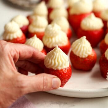 My hand reaching for a no bake strawberry cheesecake bite, from a plate of bites.