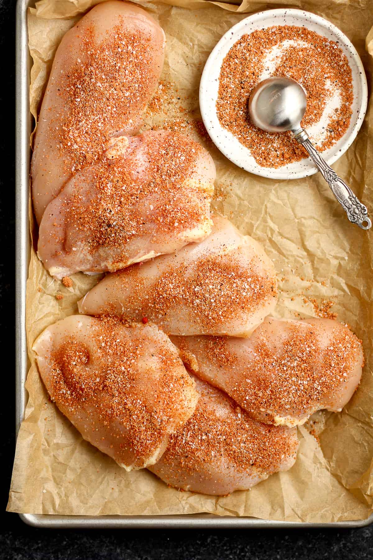 A pan of six raw chicken breasts with a small plate of seasoning.