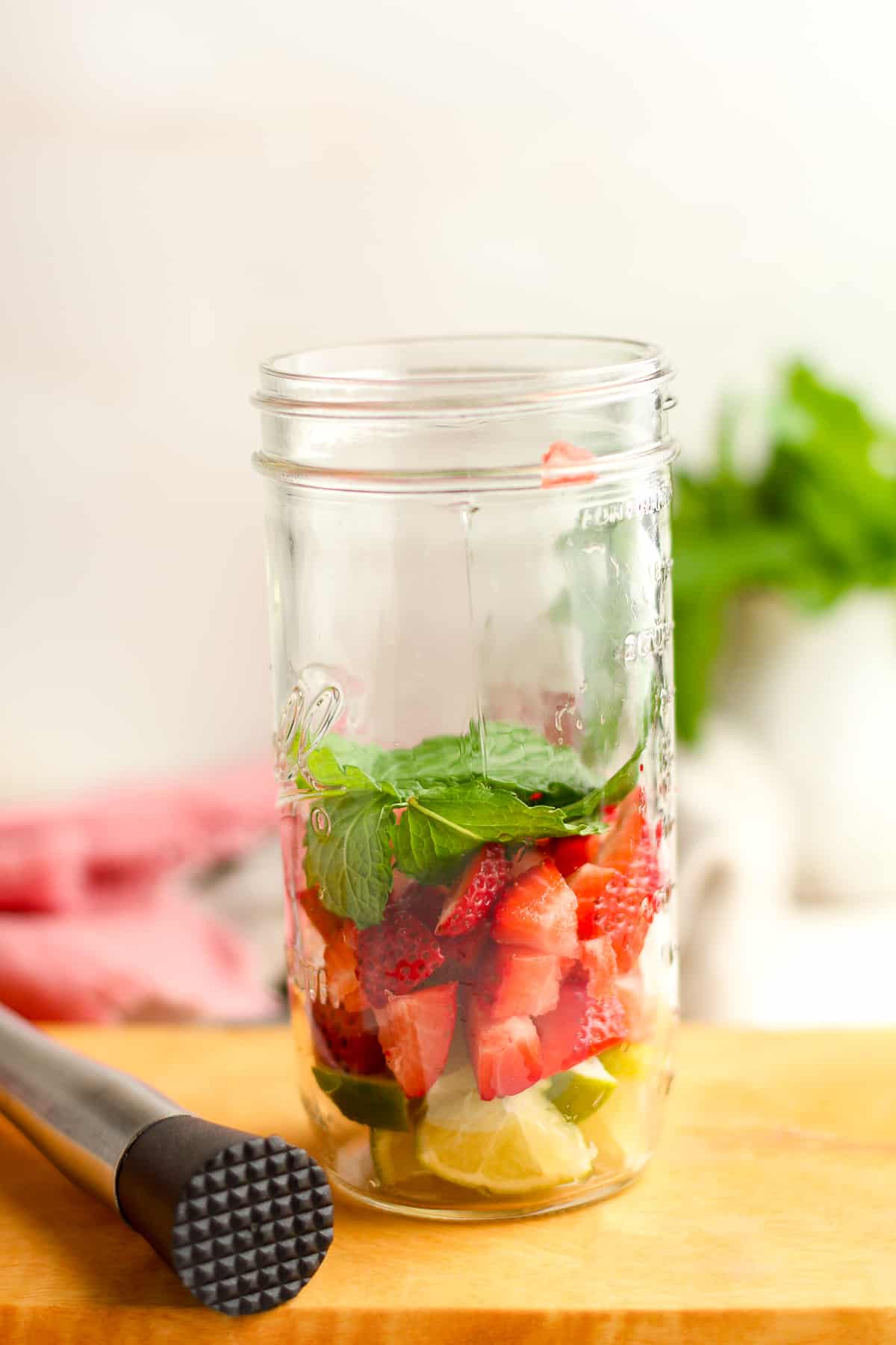 Side shot of a jar of the limes, strawberries, and mint, with a muddle next to it.