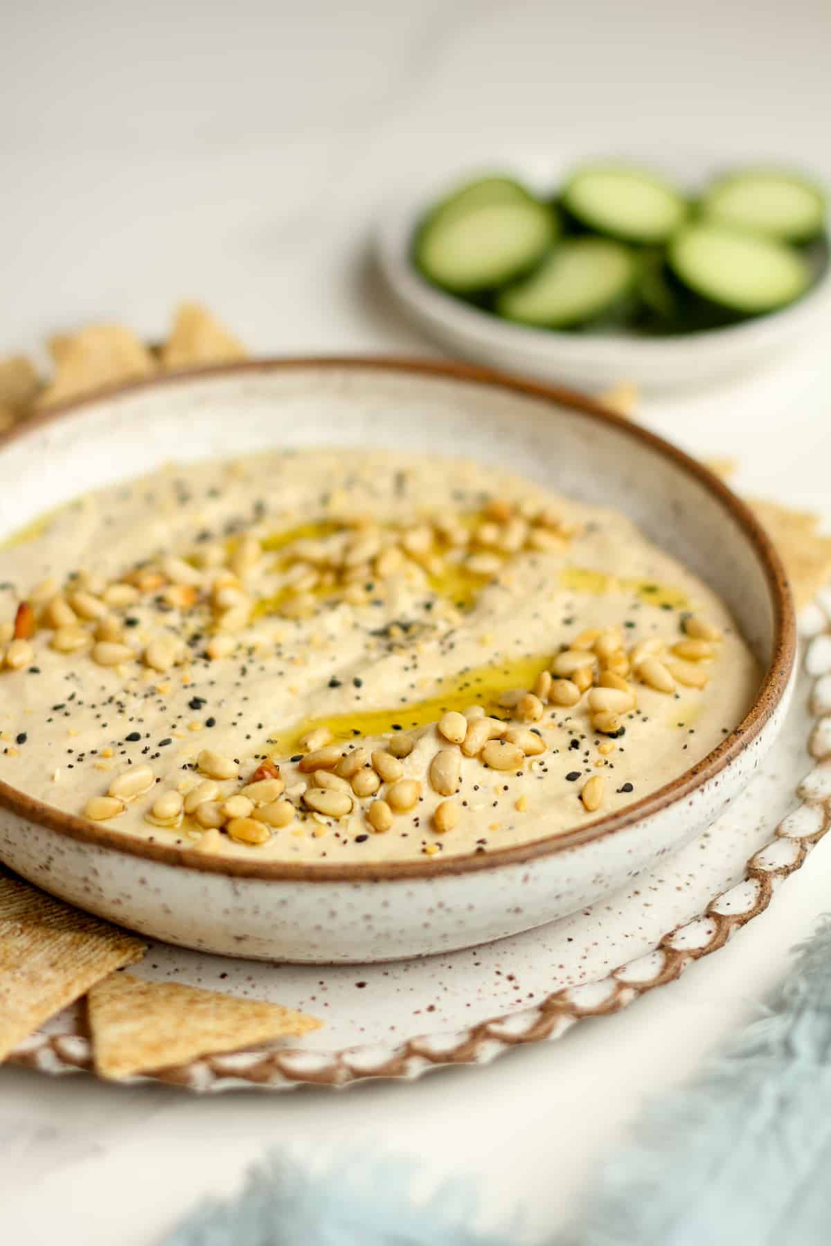 Side view of a bowl of hummus with pine nuts and crackers and cucumbers.