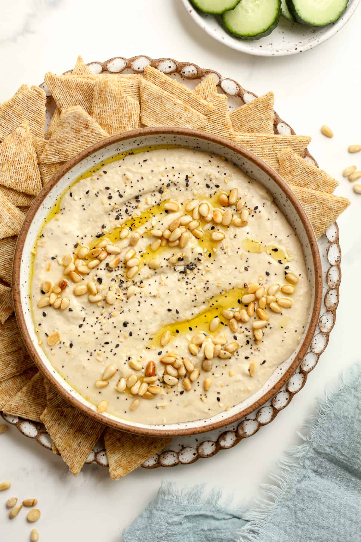 Overhead shot of a bowl of creamy hummus with toasted pine nuts with everything bagel seasoning, with crackers.