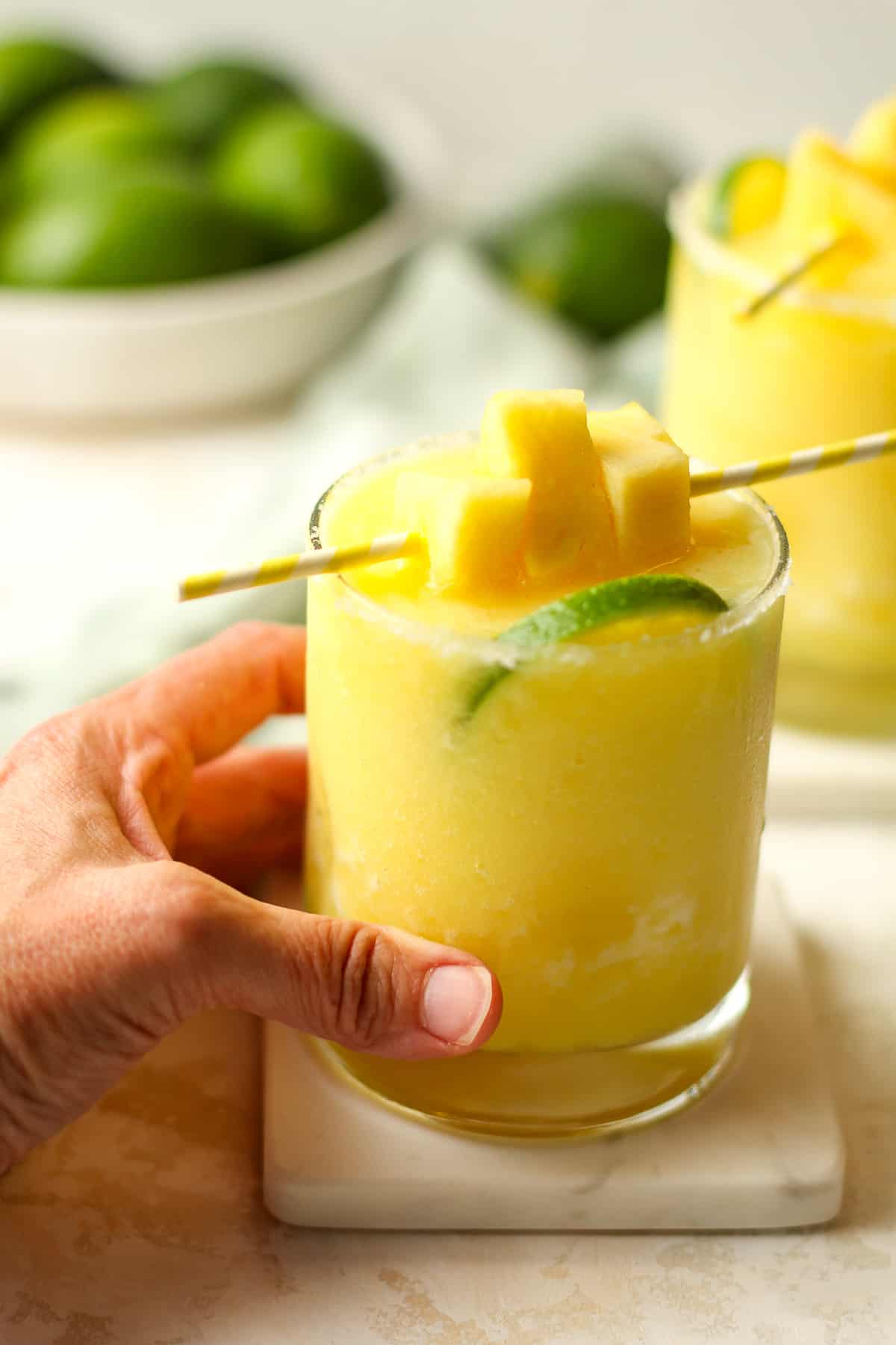 My hand on a glass of frozen pineapple margaritas with a garnish of pineapple chunks.