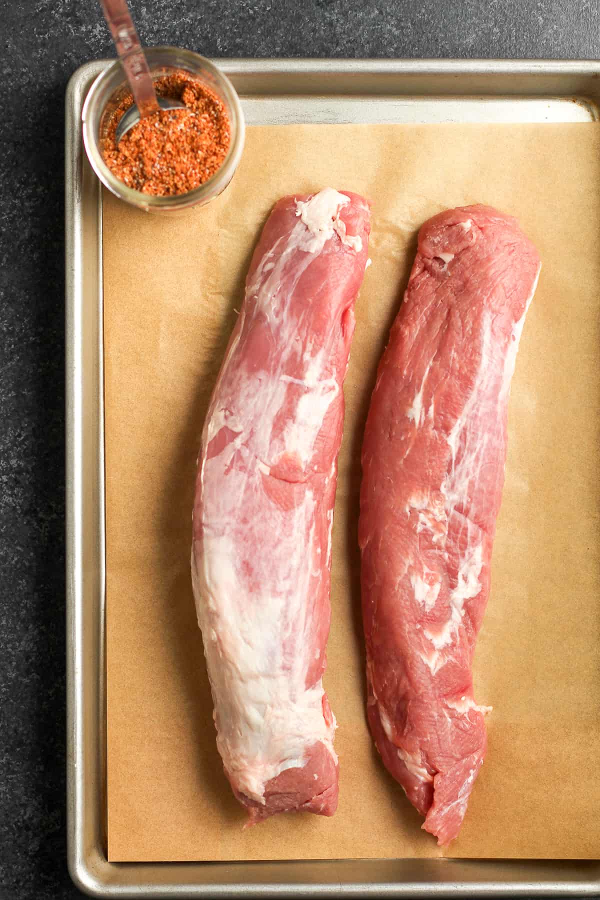 Two raw pork tenderloins on a baking sheet, with a jar of seasoning.