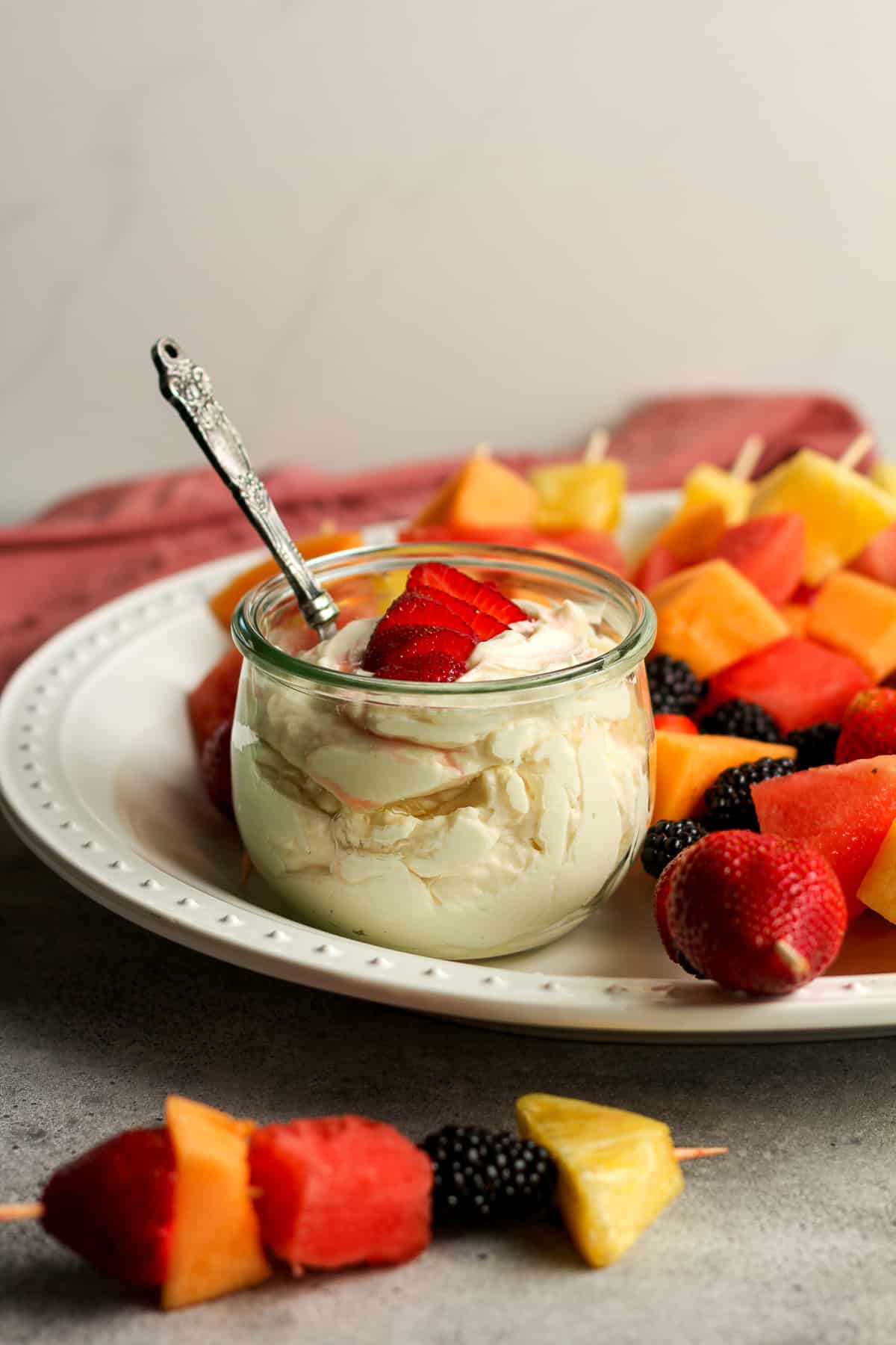 Side view of a jar of fruit dip with some fruit kabobs on a large platter.