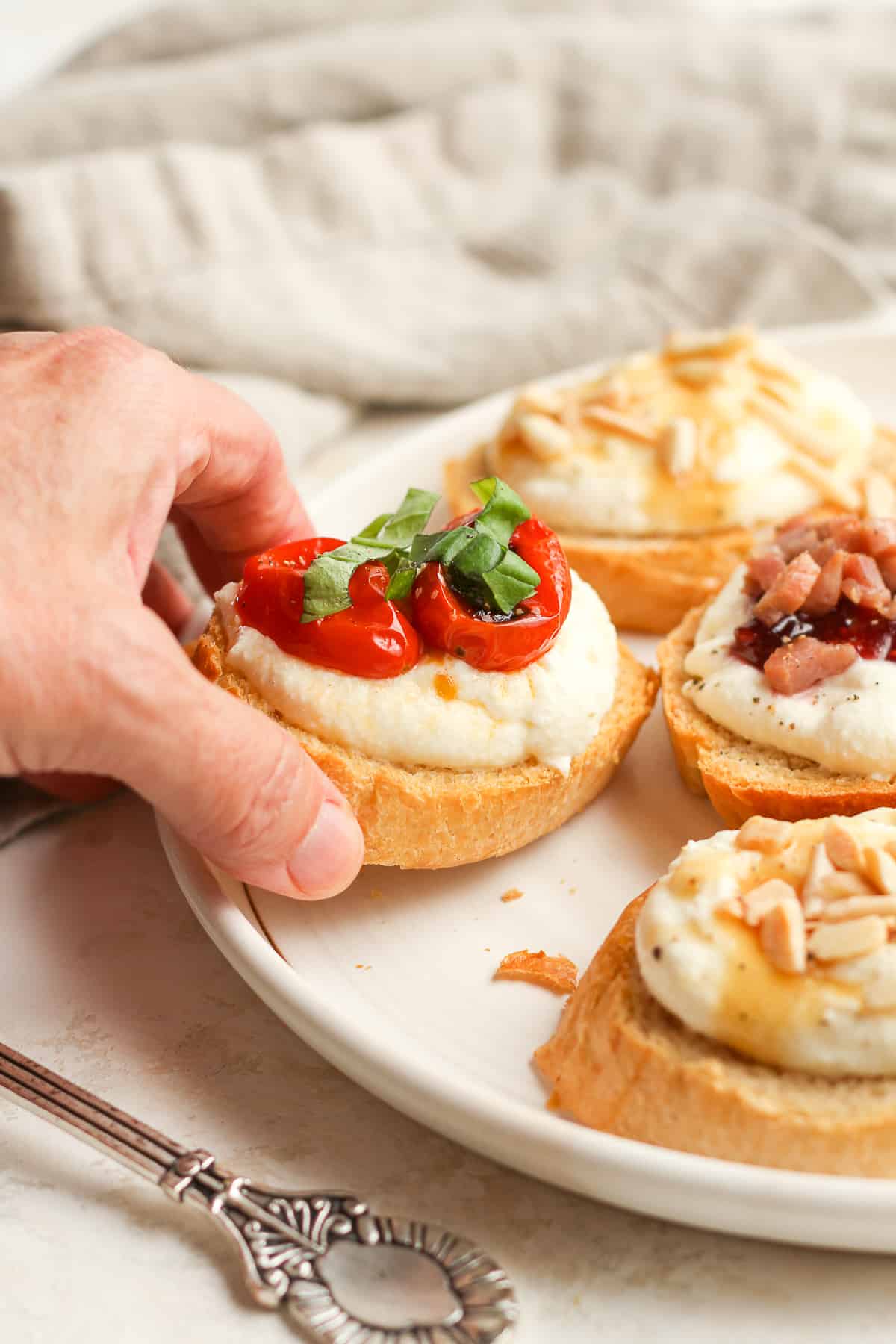 A hand reaching for a ricotta crostini on a plate of other crostini.